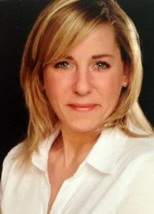Eine Frau mit einem Lächeln auf dem Gesicht. Sie hat blonde Haar und trägt eine weiße Bluse.